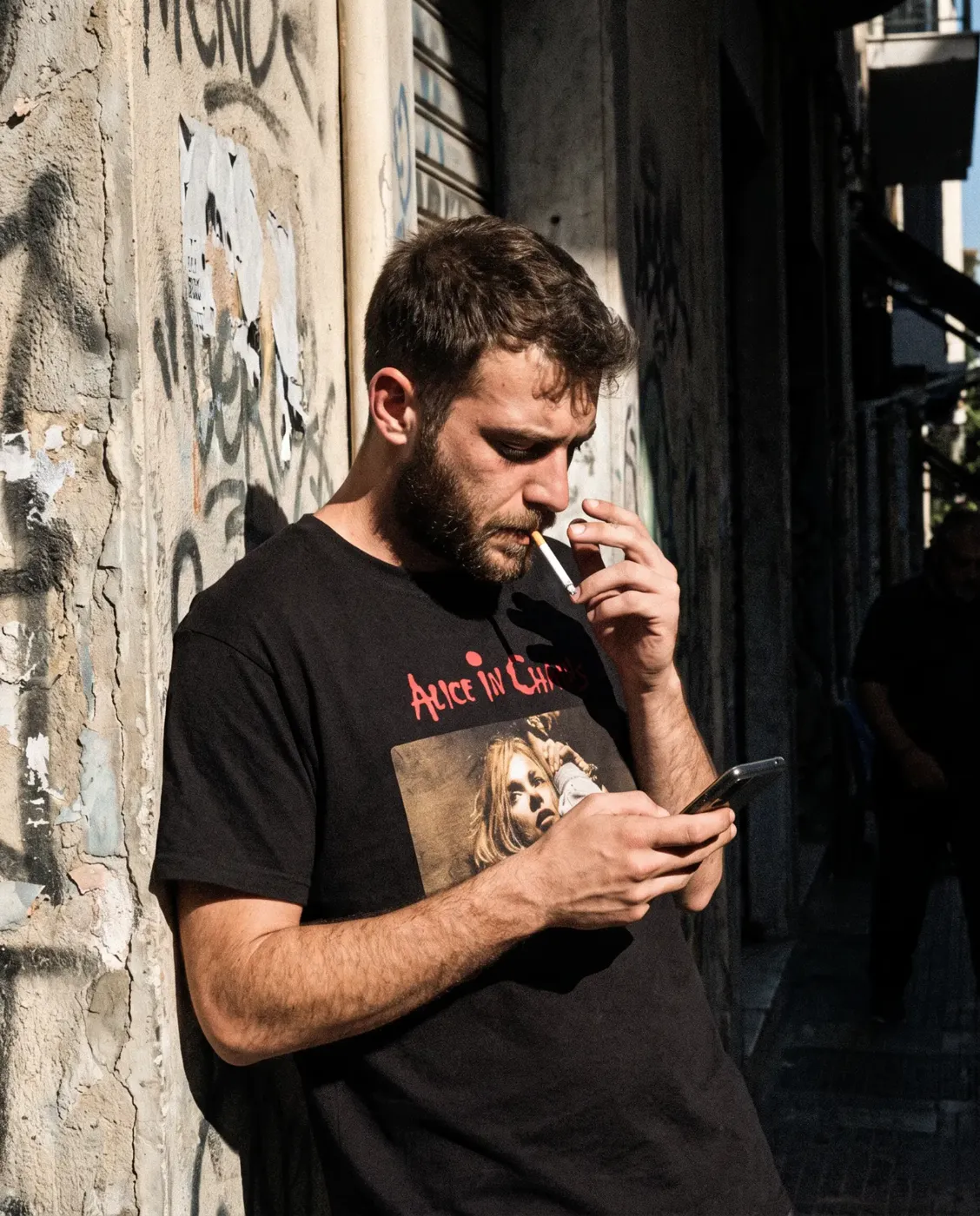 Άντρας με μαύρο t-shirt με στάμπα συγκροτήματος κοιτάζει το κινητό του σε αθηναϊκό σοκάκι με γκράφιτι